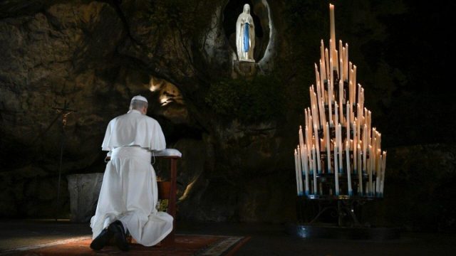 Pope Leo XIV in white kneeling in prayer before the Grotto of Massabielle at the shrine of Lourdes