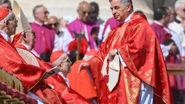Cardinal Angelo Becciu in black clerical dress with red trim, seated beside his lawyer